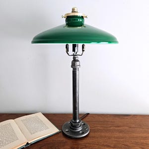 May include: A green metal lampshade with a brass top sits atop a black metal pipe lamp base. The lamp is on a wooden table with a book.