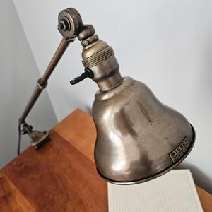 May include: A metallic industrial task lamp with an adjustable articulated arm and a clamp base. The bell-shaped shade features a label reading MICRON MACHINE CORP. BROOKLYN N.Y. and includes a black turn-key switch on the socket housing.