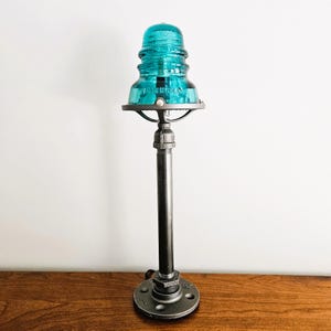 Może przedstawiać: Lampa z izolatora szklanego w kolorze turkusowym z czarną metalową podstawą i okrągłym metalowym kloszem. Lampa stoi na drewnianej powierzchni.