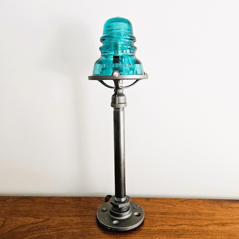 Może przedstawiać: Lampa z izolatora szklanego w kolorze turkusowym z czarną metalową podstawą i okrągłym metalowym kloszem. Lampa stoi na drewnianej powierzchni.