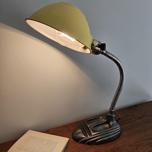 Puede incluir: Una lámpara de escritorio con una pantalla en forma de cúpula de color amarillo pálido, un cuello de metal flexible de tono plateado y una base pesada de color gris oscuro con diseño escalonado. Incluye una bombilla visible y un interruptor negro.