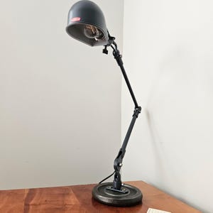 May include: A matte black desk lamp with an adjustable articulated arm and a dome-shaped shade. The lamp features a round, heavy-duty base and a visible light bulb inside the shade, which displays a small red rectangular logo label.
