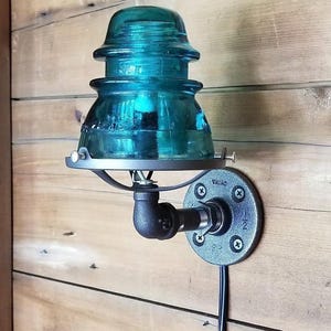 May include: A teal blue glass insulator light fixture mounted on a black metal pipe wall sconce. The sconce has a round metal plate with the word "WIRING" printed on it.