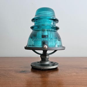 Pu&ograve; includere: Una lampada con isolatore in vetro color turchese con base e hardware in metallo nero. La lampada presenta un design vintage con la scritta "HEMINGRAY - 42" visibile sul vetro. La lampada &egrave; esposta su una superficie di legno.