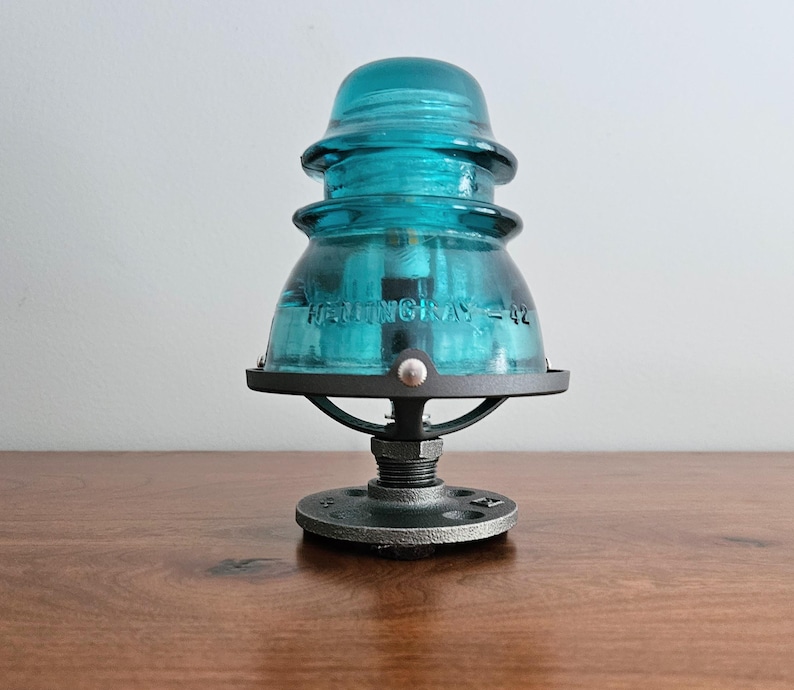 Pu&ograve; includere: Una lampada con isolatore in vetro color turchese con base e hardware in metallo nero. La lampada presenta un design vintage con la scritta "HEMINGRAY - 42" visibile sul vetro. La lampada &egrave; esposta su una superficie di legno.