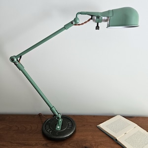 May include: A sage green adjustable desk lamp with a multi-jointed arm and a dome-shaped shade. The lamp base is a heavy, circular black metal weight plate with the text York Barbell embossed on the surface. A brown twisted fabric cord is visible.