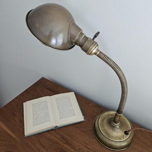 Puede incluir: Una lámpara de escritorio de latón de estilo vintage con una pantalla de cúpula redondeada, un brazo flexible de cuello de cisne y una base circular. La lámpara cuenta con un pequeño interruptor de llave en el casquillo. Un libro abierto está al lado.