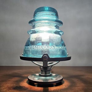 Pu&ograve; includere: Una lampada da tavolo unica con un isolatore in vetro blu vintage con la scritta "HEMINGRAY = 42". La lampada ha una base in metallo ed emette una luce calda, creando un'estetica rustica.