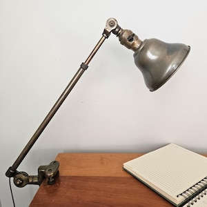 May include: Vintage brass desk lamp with an adjustable arm and bell-shaped shade. The lamp is clamped to a wooden desk, next to a spiral-bound notebook. The lamp's finish has a worn, antique appearance.