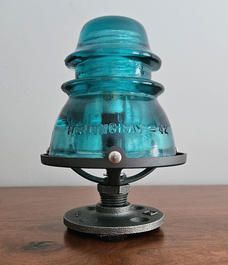 Pu&ograve; includere: Una lampada con isolatore in vetro Hemingray-42 color turchese. Il vetro &egrave; montato su una base in metallo nero con un piede rotondo in stile industriale. Il testo "HEMINGRAY = 42" &egrave; visibile sul vetro. Un oggetto decorativo unico.