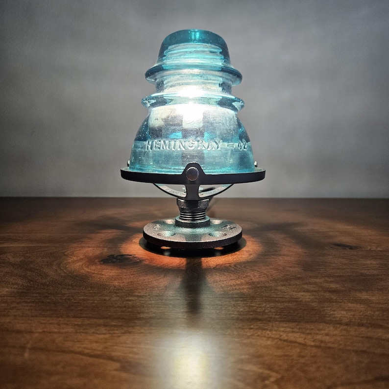 Pu&ograve; includere: Una lampada da tavolo unica con un isolatore in vetro blu vintage con la scritta "HEMINGRAY - 02". La lampada ha una base in metallo e emette una luce calda. Un elemento decorativo.