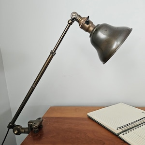 May include: An adjustable brass desk lamp with a clamp base, a long articulated arm, and a bell-shaped metal shade. The lamp features a dark metallic finish, a rotating joint at the top, and a black turn-knob switch located on the socket.
