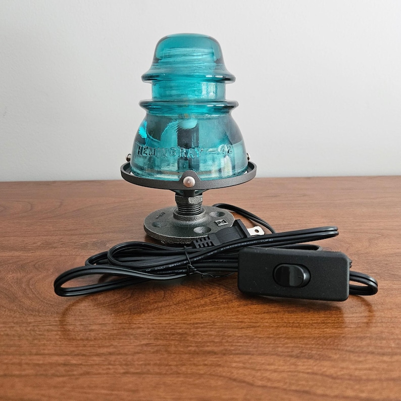 Pu&ograve; includere: Una lampada in vetro turchese con base in metallo nero e cavo nero. La lampada ha un design unico e vintage, con la scritta "HEMINGRAY - 42" visibile sul vetro. La lampada si accende con un interruttore nero.