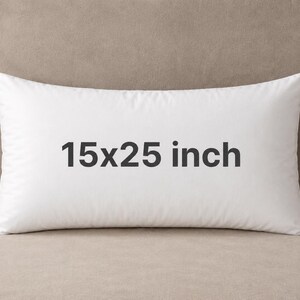 May include: A rectangular white pillow featuring the text 15x25 inch printed in dark gray on the front. The pillow has a smooth fabric surface and a small dark blue tag attached to the bottom right corner, indicating its size and construction.
