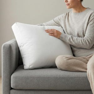 May include: A large, square white pillow rests on a gray sofa. The pillow is held by a person wearing a light gray sweater and beige pants. The sofa is in a room with a neutral color palette.