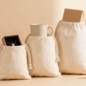 以下が含まれることがあります： さまざまなサイズの3つのクリーム色の巾着袋。それぞれに異なるアイテムが入っています。1つは小さな箱に入ったネックレス、もう1つはベージュのマグカップ、そして最大のものは小さな茶色の箱です。近くには緑の小枝があります。