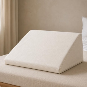 Peut inclure: Un oreiller en forme de coin avec une housse en tissu blanc cassé lisse. L'oreiller présente un profil triangulaire avec une base plate et une surface supérieure inclinée, conçu pour offrir un support incliné pour se reposer ou s'asseoir.