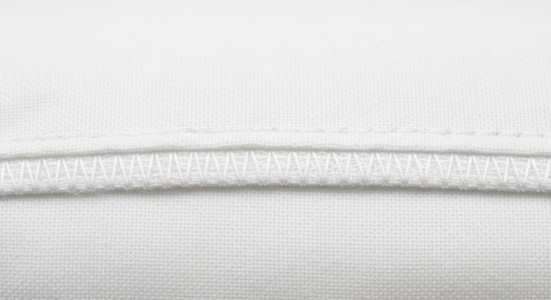 May include: Close-up of white fabric with two rows of stitching. The top row is a straight stitch, and the bottom row is a zigzag stitch. The fabric appears to be a tightly woven cotton or linen.
