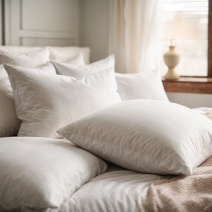 Op de afbeelding: Een close-up van een bed met meerdere witte kussens die in lagen zijn gerangschikt. De kussens zijn zacht en pluche, en het bed is gemaakt met frisse witte lakens. Een lichtbruine deken is over de onderkant van het bed gelegd.