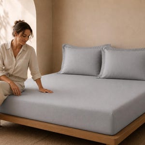 May include: A light gray fitted sheet covering a mattress on a wooden bed frame, accompanied by two matching light gray pillowcases with wide borders. The bedding set features a smooth, solid-colored fabric texture across the entire surface.
