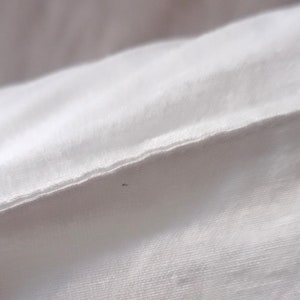 May include: Close-up of white fabric, possibly a sheet or pillowcase, with a visible seam. The fabric appears to be smooth and tightly woven, with subtle texture. The lighting is soft, highlighting the folds and details of the material.