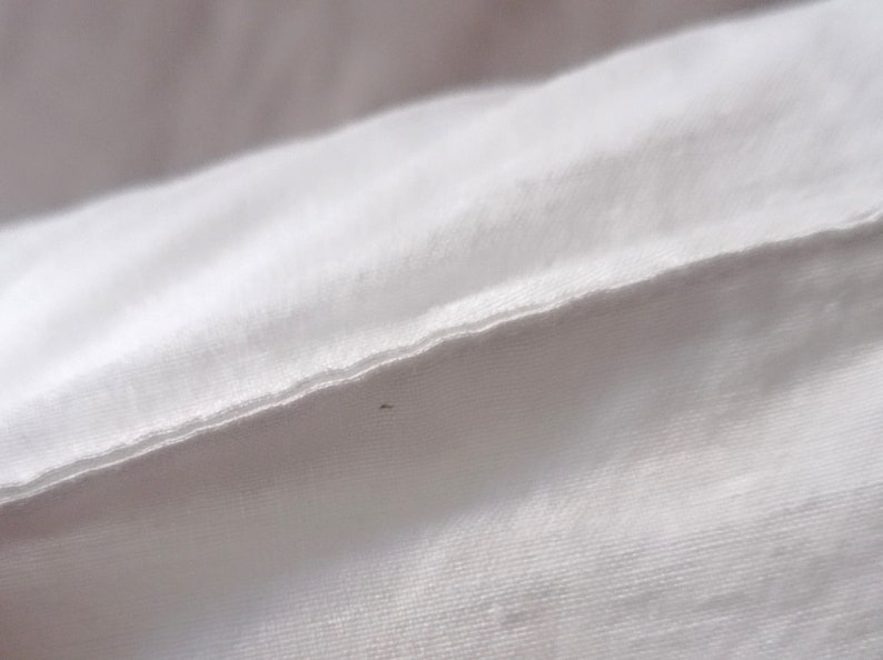 May include: Close-up of white fabric, possibly a sheet or pillowcase, with a visible seam. The fabric appears to be smooth and tightly woven, with subtle texture. The lighting is soft, highlighting the folds and details of the material.