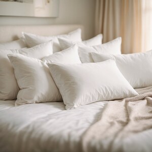 Puede incluir: Una cama blanca con un edredón blanco y una pila de almohadas blancas. Una manta marrón claro está drapeada sobre las almohadas.