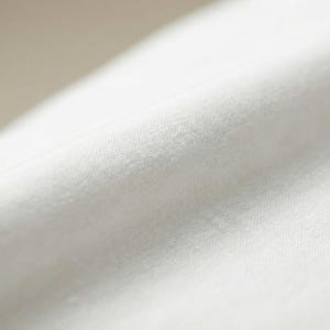 May include: Close-up of a white fabric with a soft, textured surface. The fabric appears to be tightly woven, with subtle variations in tone and texture. The image highlights the material's smooth, flowing drape.