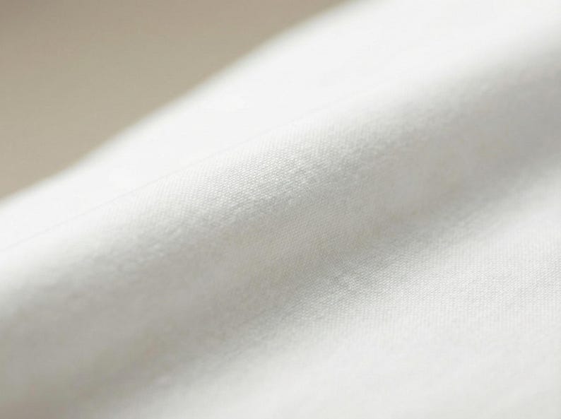 May include: Close-up of a white fabric with a soft, textured surface. The fabric appears to be tightly woven, with subtle variations in tone and texture. The image highlights the material's smooth, flowing drape.
