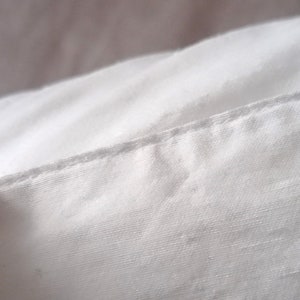 May include: Close-up of white fabric with a visible seam. The fabric appears to be a tightly woven material, possibly cotton or linen. The seam is a straight stitch, indicating a tailored construction. The lighting creates soft shadows.