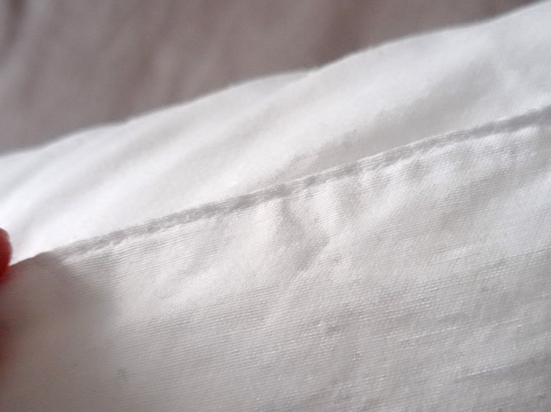 May include: Close-up of white fabric with a visible seam. The fabric appears to be a tightly woven material, possibly cotton or linen. The seam is a straight stitch, indicating a tailored construction. The lighting creates soft shadows.