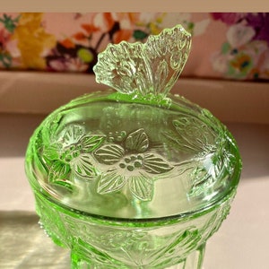 May include: A green glass trinket dish with a butterfly-shaped handle. The dish features floral and leaf patterns and sits on three small feet. The lid and base are round, and the glass has a slight glow.
