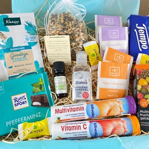 Könnte beinhalten: Eine blaue und weiße Geschenkbox gefüllt mit verschiedenen Wellness- und Gesundheitsprodukten, darunter Badekristalle, Teebeutel, Händedesinfektionsmittel, Vitaminpräparate und eine Schokoladentafel. Die Box ist mit zerkleinertem Papier gefüllt und hat eine blau-weiß gestreifte Auskleidung.