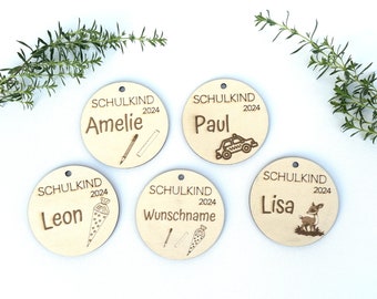 Pode incluir: Cinco ornamentos redondos de madeira com o texto "SCHULKIND 2024" e um nome em cada um. Os nomes são Amelie, Paul, Leon, Wunschname e Lisa. Cada ornamento tem um design diferente, incluindo um lápis e uma régua, um táxi, um cone escolar, um lápis e um cone escolar, e um veado.
