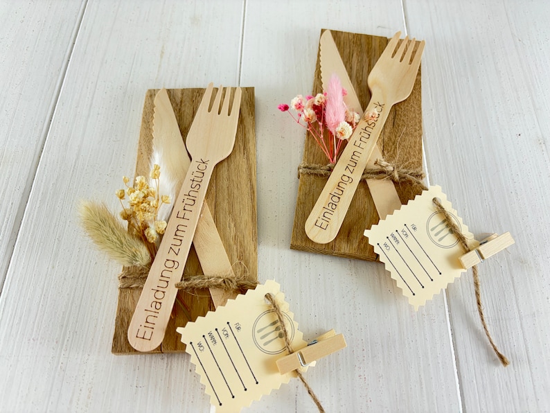 K&ouml;nnte beinhalten: Zwei Holz-Platzkarte mit je einer Gabel und einem Messer. Die Platzkarte sind mit getrockneten Blumen und Schnur verziert. Der Text auf den Platzkarte lautet "Einladung zum Fr&uuml;hst&uuml;ck".