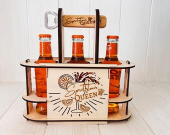 May include: A wooden carrier holding three orange-colored bottles. The carrier has a built-in bottle opener and features the text "Spritz Your Queen". The design includes a graphic of a cocktail and citrus slices.