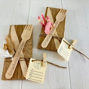 K&ouml;nnte beinhalten: Zwei Holz-Einladungen zum Grillen, jede mit einem Holz-Messer und einer Holz-Gabel, mit Schnur gebunden und mit getrockneten Blumen verziert. Die Einladungen sagen "Einladung zum Grillen".