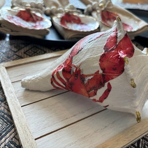 Könnte beinhalten: Eine weiße Muschel mit einem roten Hummer-Design und goldenen Akzenten. Die Muschel liegt auf einem Holztablett mit weißer Oberfläche.