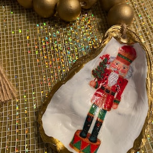 Puede incluir: Una concha de ostra blanca con un borde dorado, pintada con una figura de cascanueces. El cascanueces lleva un uniforme rojo y verde con un sombrero negro y sostiene un bastón de caramelo.