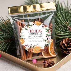 Peut inclure: Un sac transparent avec un dessus doré, rempli de bâtons de cannelle, de tranches d'orange séchées et d'anis étoilé. Une étiquette ronde indique "HOLIDAY SIMMERING POTPOURRI". Le sac est posé sur un plateau doré avec des aiguilles de pin et une pomme de pin.