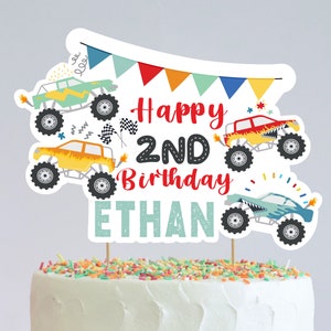 May include: Monster truck themed cake topper with colorful flags, a red "Happy 2nd Birthday" message, and the name "Ethan" in teal. Four monster trucks are featured in the design.