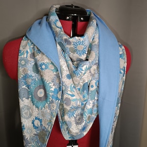 May include: A blue and white floral patterned scarf with a light blue solid fabric border. The scarf is folded in a triangular shape and draped over a mannequin.