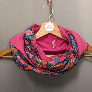 May include: An infinity scarf featuring a vibrant floral pattern with shades of pink, orange, teal, and black on one side and a solid bright pink fabric on the reverse. The scarf is draped on a wooden hanger with a Liberty Stories brand label.