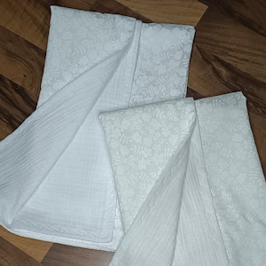 May include: Two white muslin swaddles with a delicate floral pattern. The swaddles are folded and layered on a wooden surface.