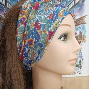 Peut inclure: Un bandeau à imprimé floral avec des fleurs rouges, bleues, jaunes et vertes. Le bandeau est porté sur une tête de mannequin avec des cheveux bruns. L'arrière-plan comprend une peinture colorée avec divers objets.