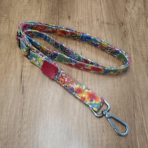 Op de afbeelding: Een kleurrijke, met bloemen bedrukte band met een zilveren metalen haak. De band is gemaakt van stof en heeft een rood leren patch.