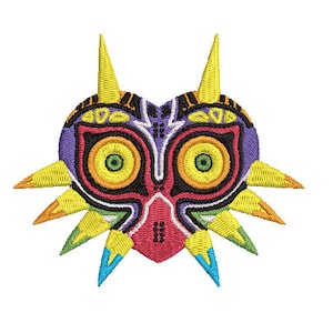 May include: A colorful embroidered patch featuring the Majora's Mask from the Legend of Zelda video game series. The mask is a heart shape with a fierce expression and sharp, pointed teeth. The mask is surrounded by a ring of yellow, green, and blue spikes.