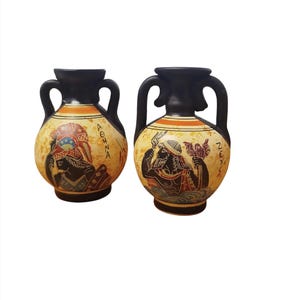 May include: Two small black ceramic vases with handles. The vases are decorated with hand-painted scenes of Greek mythology. One vase features a woman with a headdress and the word "ATHENA" written on it. The other vase features a man with a trident and the word "POSEIDON" written on it.
