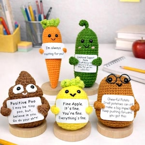 May include: A collection of handmade crochet figures, including a carrot, pickle, poop, apple, and potato, each holding a sign with an encouraging message. The figures are on wooden bases, with pencils and a notebook in the background.