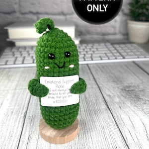 Könnte beinhalten: Ein handgehäkelter grüner Krokodilgurke mit einem lächelnden Gesicht und kleinen Armen, der ein Schild mit der Aufschrift "Emotional Support Pickle" hält. Die Gurke steht auf einem Holzsockel. Ein schwarzer Kreis mit "PDF PATTERN ONLY" befindet sich im Hintergrund.
